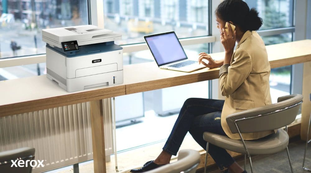 Une personne est assise à une table haute près d'une grande fenêtre, parlant au téléphone tout en travaillant sur un ordinateur portable. Une imprimante multifonction Xerox B235DNI est judicieusement placée sur la table à côté d'elle, suggérant une utilisation simple dans un espace de travail moderne.